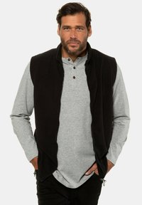 Schwarze Fleeceweste über einem grauen Langarmshirt, mit Reißverschluss, Knopf-Akzenten und Seitentaschen.