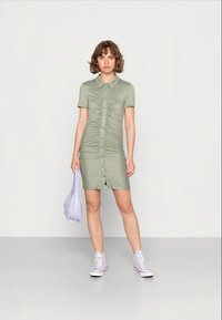 Vestido verde oliva de manga curta com botões frontais e textura franzida. Combinado com uma bolsa roxa e ténis rosa claro.