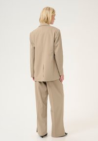 Blazer beige ajusté avec une fente unique, associé à un pantalon à jambes larges. Tissu lisse, forme structurée et sans motifs visibles.