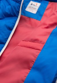 Close-up van een blauwe en rode jas met een zichtbaar label op het bovenste blauwe gedeelte en gedetailleerde stiksels op de rode stof.