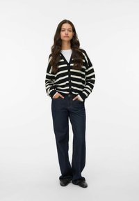 Cardigan rayé noir et blanc avec un col en V, fermeture à boutons et manches longues, associé à un jean large en denim foncé et des chaussures noires.