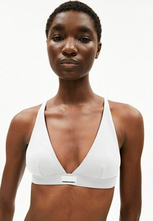 Femme aux cheveux courts portant un haut de soutien-gorge blanc sans manches avec l’étiquette « ARMEDANGELS », debout sur un fond blanc uni.