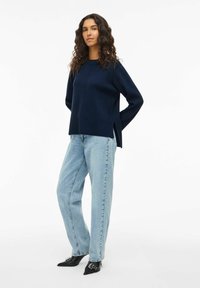 Pull en tricot bleu marine à manches longues, avec des fentes latérales, associé à un jean à pattes d'éléphant bleu clair et des escarpins noirs à bout pointu avec des accents décoratifs.