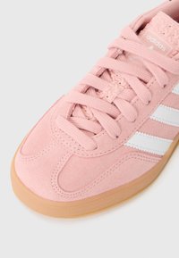 Sapatilha de camurça rosa com design de três riscas brancas, bico arredondado e língua texturada. Apresenta uma sola de borracha cor de nozes e atacadores planos.