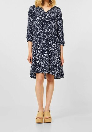 Vrouw draagt een marineblauwe jurk met witte polkadots en beige plateauzandalen, staand tegen een effen lichte achtergrond.