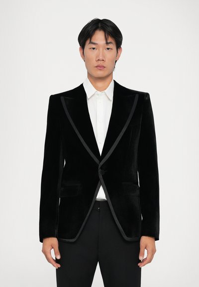 Veste de smoking en velours noir avec des garnitures en satin et des revers crantés, dotée d'une fermeture à un bouton et de deux poches avant. Chemise blanche à manches longues en dessous.
