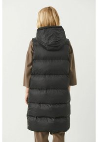 Persoon in een lange zwarte gewatteerde mouwloze capuchonvest over een bruin lange mouwen top, staand met de rug naar de camera tegen een effen achtergrond.