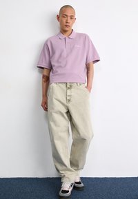 Helllila Kurzarm-Polohemd mit weicher Textur, kombiniert mit locker sitzenden beigen Jeans und schwarzen Schuhen mit Pinkakzenten.