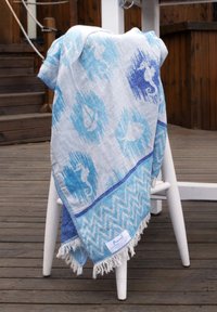 Light blue and white patterned throw with seahorse, sailboat, and fish designs draped over a white wooden chair on wooden decking.