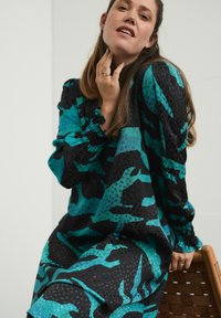 Šaty v odtieňoch teal a čiernej s rozmanitými textúrami, dlhými rukávmi a zberanými manžetami, s voľnejším strihom.