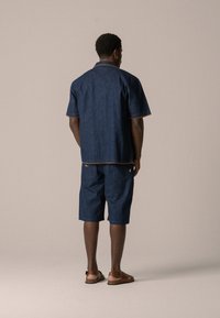 Navy denim short-sleeve shirt and shorts set with orange stitching details and two rear pockets, paired with brown sandals.