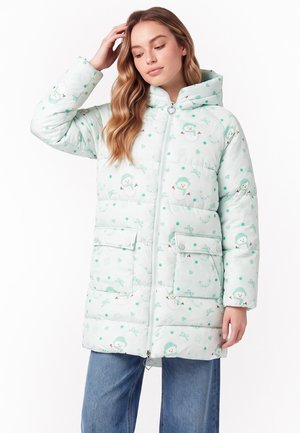Frau trägt eine hellblaue Steppjacke mit Kapuze, verziert mit Cartoon-Schneemännern und grünen Punkten, steht mit einer Hand im Haar.