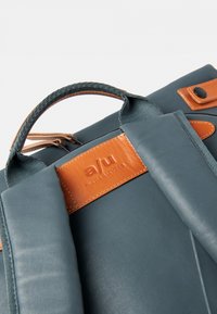 Zaino grigio in tessuto con dettagli in pelle marrone. Presenta una maniglia superiore, spallacci imbottiti e un'etichetta con il nome del marchio.