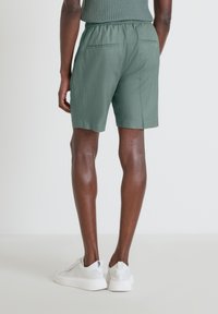 Pantaloni corti verdi in cotone con vita elastica, caratterizzati da una texture liscia e due tasche posteriori, abbinati a sneaker bianche.