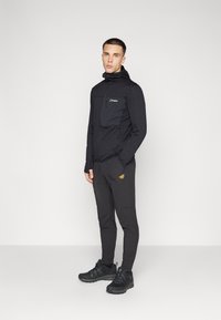 Schwarze Zip-Jacke mit einer Brusttasche und strukturiertem Stoff, kombiniert mit schwarzen Hosen mit einem Logo und akzentuiertem Schuhwerk.