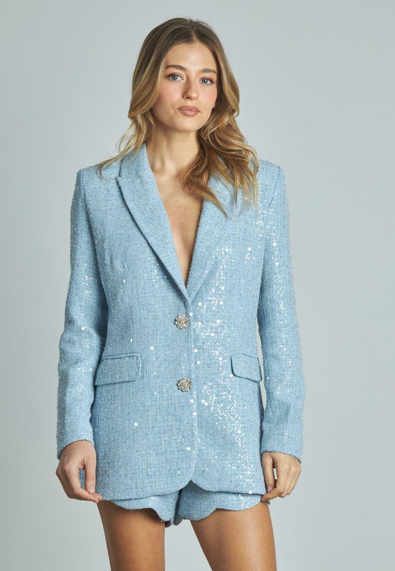 Blazer azzurro chiaro con paillettes, scollatura a V profonda, due tasche anteriori e bottoni decorativi. Il tessuto strutturato crea un effetto scintillante.