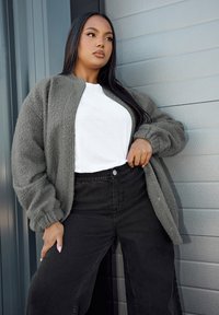 Grå fluffig jacka med krage som bärs över en vit t-shirt, ihopparad med högmidjade svarta byxor. Slät textur med avslappnad passform.