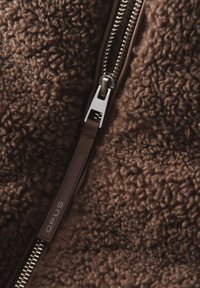 Braunes Fleece-Material mit einer strukturierten, plüschigen Oberfläche; ausgestattet mit einem silbernen Reißverschluss und einem braunen Zugband, das mit "OPUS" beschriftet ist.