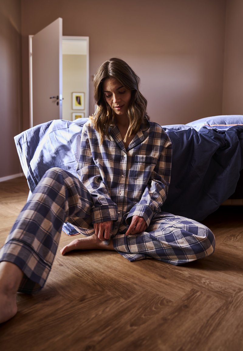 Blå og beige ternede flannelpyjamaser med knaplukning foran og løst siddende bukser, båret mens man sidder på et trægulv.