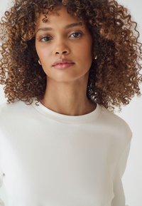 Felpa bianca con collo rotondo e maniche lunghe, caratterizzata da una texture liscia. Il modello ha capelli ricci e trucco naturale.