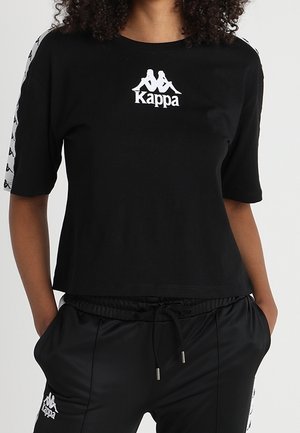 Persoon draagt een zwart Kappa T-shirt met korte mouwen en logo en bijpassende zwarte broek met trekkoord, handen in de zakken, tegen een effen achtergrond.