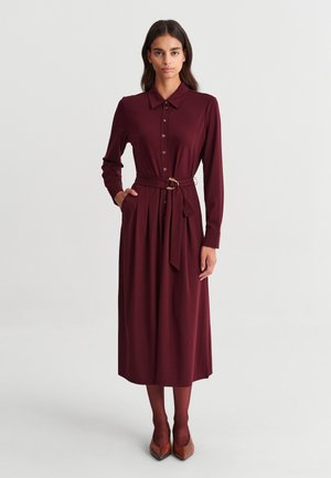 Femme vêtue d'une robe bordeaux à manches longues boutonnée, avec une ceinture, des collants marron et des chaussures marron à bout pointu, debout devant un fond clair uni.