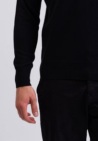 Schwarzer gestrickter Pullover mit Rundhalsausschnitt und gerippten Bündchen, kombiniert mit dunklen Stoffhosen. Die Hand ist natürlich an der Seite positioniert.