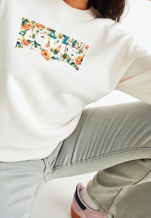 Personne portant un sweat-shirt blanc avec un logo floral "Levi's", un jean gris clair et des baskets roses assise avec un genou plié.