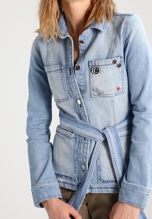 Lichtblauwe spijkerjack met voorzakken, knoopsluitingen, riem gestrikt in de taille en decoratieve pins op het linkerborstzakje.