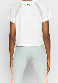 Camiseta blanca recortada con paneles de malla en los hombros. Presenta un pequeño logo negro en la parte trasera, combinada con leggings verde claro con detalles en coral.