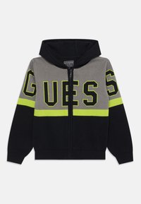 Veste à capuche zippée en gris, noir et jaune fluorescent. Caractérisée par un grand logo "GUESS" et un blocage de couleurs contrasté. Matière en tricot douce.
