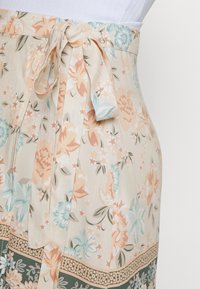 Saia envolvente floral em creme com flores laranja, verde-água e verdes. Apresenta uma cintura atada e pregas de lado. Tecido macio e leve.