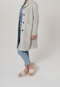 Abrigo de houndstooth en tonos gris y blanco con solapa, dos bolsillos frontales y tres botones, combinado con unos vaqueros azul claro y sandalias beige.