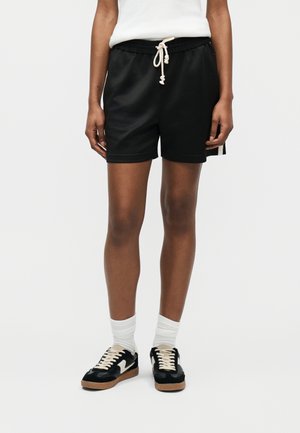 Person iført sorte træningsshorts med hvid snøre, hvide mellemlange sokker og sorte og hvide sneakers, stående mod en ensfarvet baggrund.