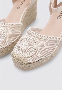 Sandalias de cuña beige con parte superior de tela trenzada, bordado intrincado y suela de yute texturizada. Correa ajustable en el tobillo para un mejor ajuste.