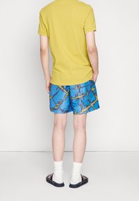 Polo en coton jaune, shorts bleus avec un motif abstrait audacieux en vert et violet, chaussettes blanches et sandales noires, vu de dos.