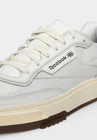 Λευκό αθλητικό παπούτσι Reebok με κρεμ κορδόνια, εμπορικό σήμα και σημαία Union Jack στο πλάι, μπεζ ενδιάμεση σόλα και καφέ λαστιχένια εξωτερική σόλα.