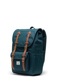 Mörk teal ryggsäck tillverkad av tyg med bruna läderremmar och guldfärgad hårdvara, med ett huvudflap och en framficka med en logotyp patch.