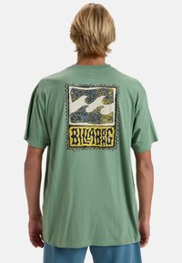 Zelené bavlněné tričko s velkým, vícolor grafickým designem na zadní straně, zobrazující vlnové vzory a slovo "Billabong" tučným písmem.