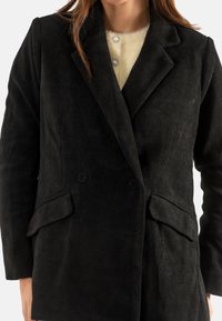 Cappotto doppiopetto in corduroy nero con ampi bavero, tasche a patta, indossato sopra un maglione crema con bottoni decorativi in perle.
