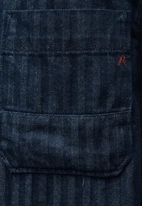 Denimstoff med mørkeblå fiskebensmønster som har en rektangulær lomme og liten rød brodert detalj på øvre høyre side.