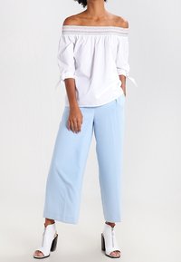 Donna che indossa una blusa bianca a spalle scoperte con maniche annodate, pantaloni larghi blu chiaro e stivaletti con tacco aperti bianchi.