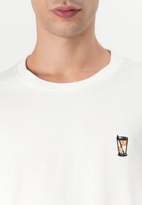 Hvit bomulls t-skjorte med et lite brodert glassdesign med appelsiner og et sugerør på venstre brystområde. Enkel rund hals.