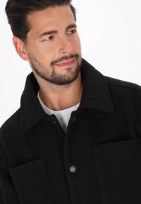 Bärtiger Mann, der einen schwarzen Knopf-Mantel über einem weißen Hemd trägt und mit einem leichten Lächeln zur Seite schaut.