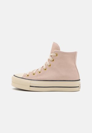 Zapatillas altas en ante rosa claro con ojetes dorados, una suela de goma beige y ribete negro alrededor de la suela. Diseño clásico de capuchón en la punta.