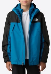 Blauw-zwarte capuchonjas met ritssluiting aan de voorkant, twee zijzakken en een logo op de schouder, met een zachte voering aan de binnenkant.