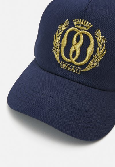 Tume sinine pesapalli kork, mis on valmistatud kangast ja millel on kuldne brodeeritud embleem, millel on põimunud ringid ja kroon, ning detailne niiditöö ääre peal.