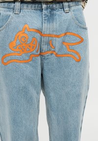 Ανοιχτό μπλε τζιν παντελόνι με πορτοκαλί περίγραμμα γραφικού ενός καρτούν σκύλου που τρέχει στην μπροστινή πάνω περιοχή του μηρού.