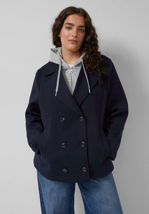 Junge Frau mit lockigem Haar, die einen marineblauen zweireihigen Mantel über einem grauen Kapuzenpullover mit Reißverschluss und blauer Jeans trägt, mit den Händen in den Taschen stehend.
