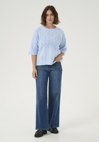 Gaiši zils adīts džemperis ar teksturētu rakstu, apaļu kakla izgriezumu un īsām piedurknēm, kopā ar platām tumši zilām biksēm un melniem apaviem.